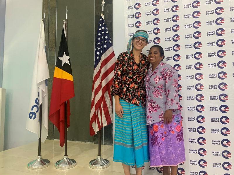 Two people smile, arm-in-arm, wearing colorful garb in front of a Peace Corps banner